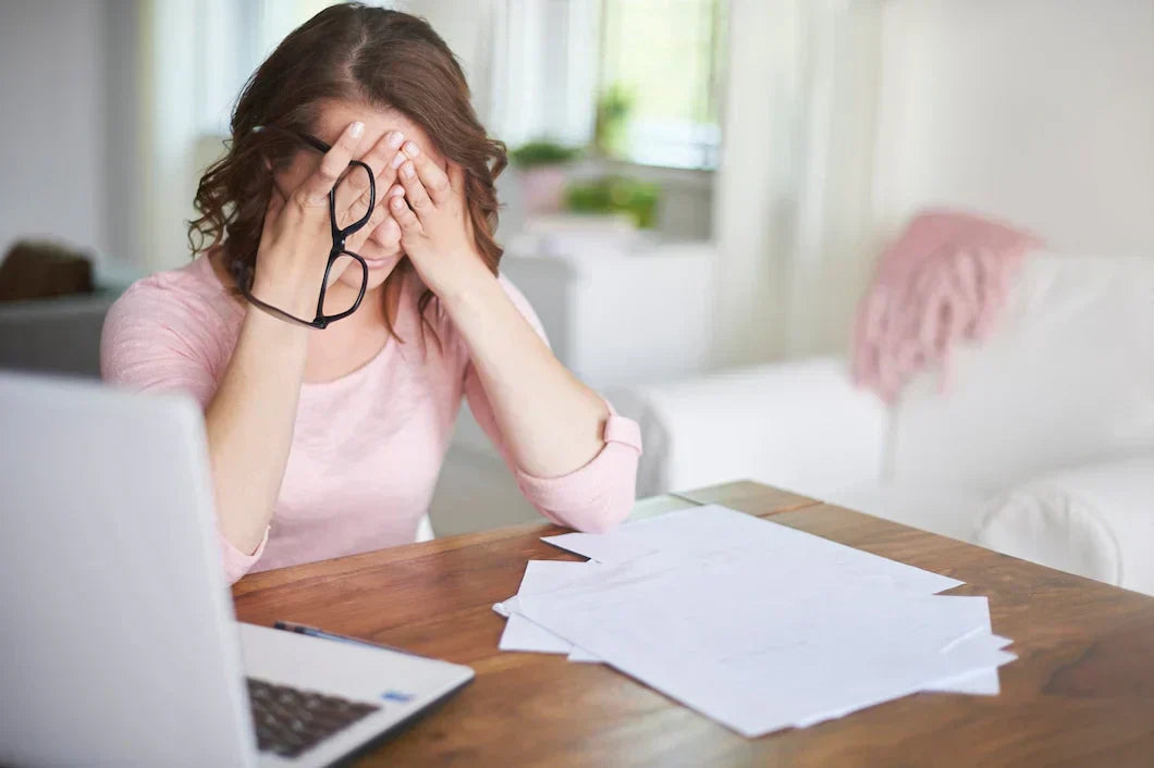 A stressed woman at her desk holds her head in her hands, surrounded by paperwork and a laptop, representing the impact of chronic stress on cardiovascular health.